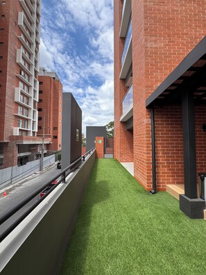 A narrow pathway with artificial grass, flanked by red brick buildings with balconies and a clear sky above. - Aurea Luxury Apartment Z10 (Guatemala)