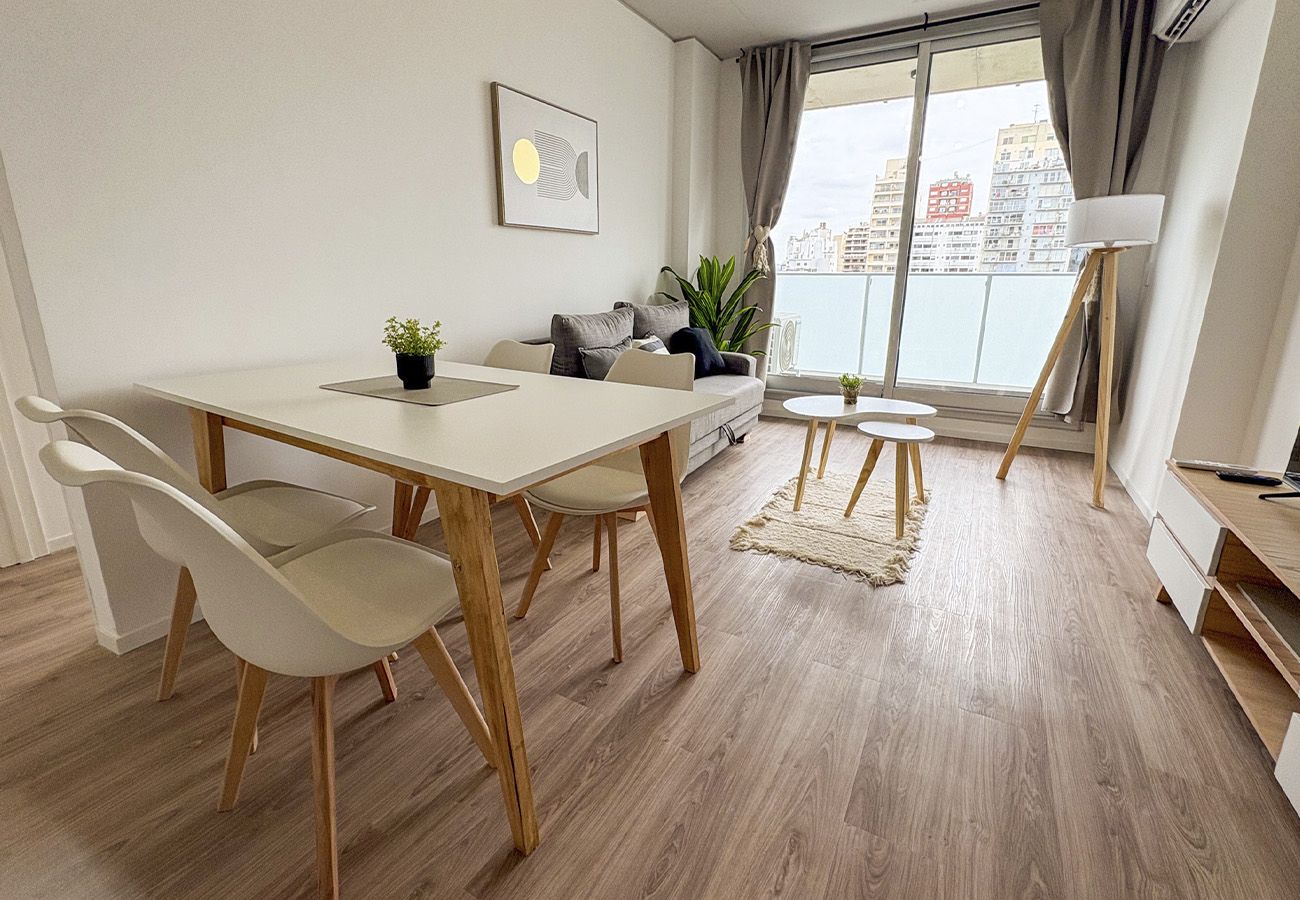 Una sala de estar moderna con una mesa de comedor, sillas, un sofá, una pequeña mesita auxiliar, una lámpara de pie y vistas de la ciudad a través de amplios ventanales.