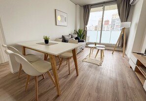 Una sala de estar moderna con una mesa de comedor, sillas, un sofá, una pequeña mesita auxiliar, una lámpara de pie y vistas de la ciudad a través de amplios ventanales.