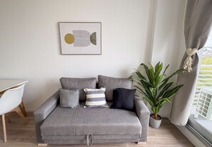 Un sofá gris con varios cojines, una obra de arte abstracta enmarcada, una planta en maceta y una silla blanca.