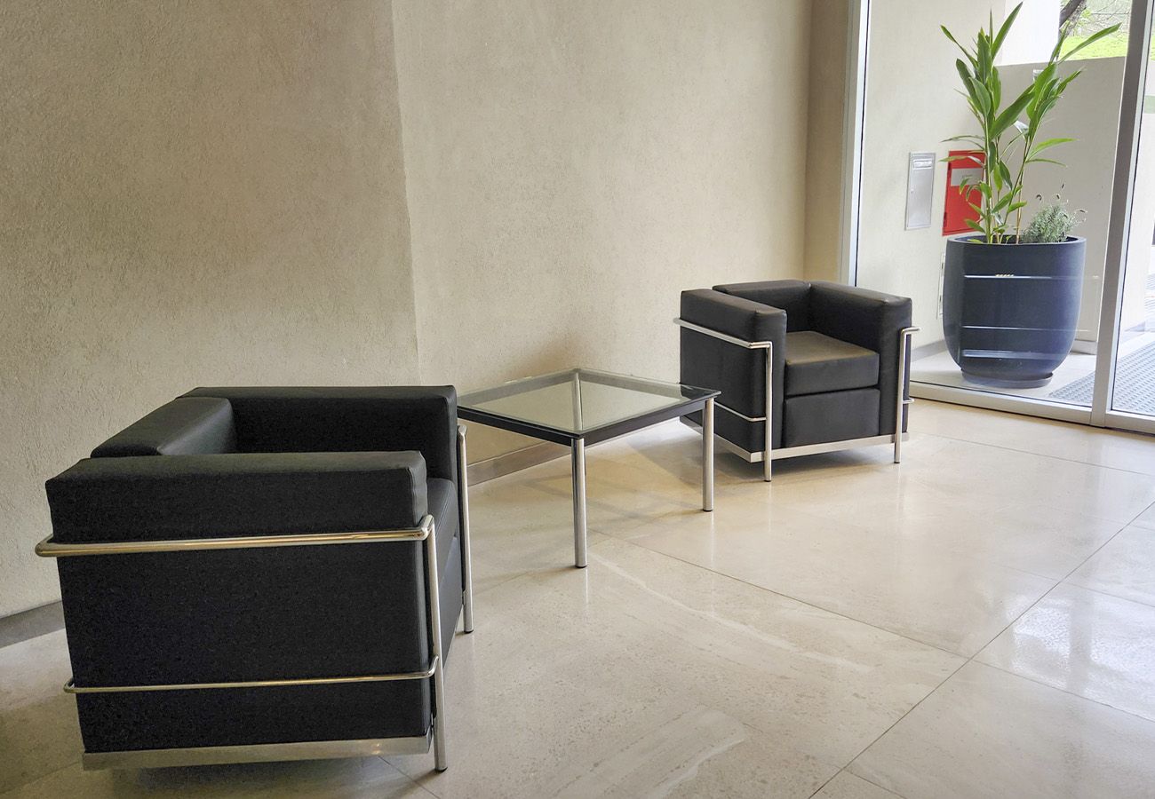 Eine moderne Lobby mit zwei schwarzen Sesseln, einem Glastisch, einer großen Topfpflanze und einem Feuerlöscher an der Wand.