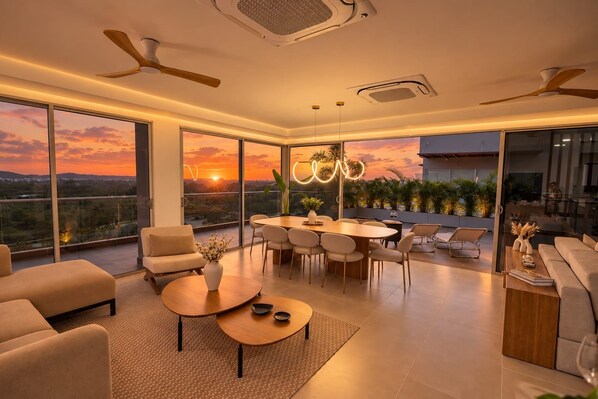 Ein modernes Wohnzimmer mit Essbereich, großen Schiebeglastüren, Deckenventilatoren und Blick auf einen Sonnenuntergang.
