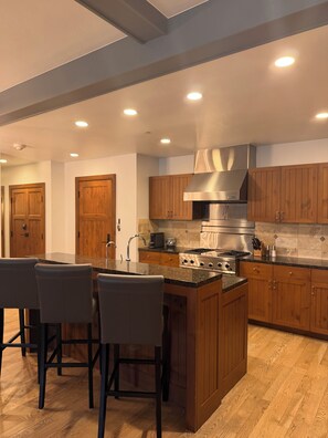 A modern kitchen with wooden cabinets, a central island with bar stools, a stainless steel range hood, and recessed ceiling lighting. - Snow Cloud Slope Side Dream
3 bedroom/3 1/2 bath ski in/ski out (Avon)