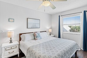 A bedroom with a bed, bedside lamps, a ceiling fan, a window with curtains, and a wall with a framed picture. - Portside Perch 2-Condo Coastal Retreat • Pool • Hot Tub • Game Room • Steps to the Beach (Indian Rocks Beach)