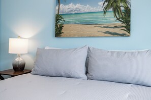 A bed with two pillows, a nightstand with a lamp, and a painting of a beach scene on the wall. - Spanish Villas #58 - Block From The Beach (Indian Rocks Beach)