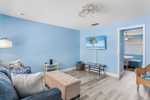 A room with blue walls, a wooden floor, a sofa, a table, a bench, a TV, and a ceiling fan. - Spanish Villas #58 - Block From The Beach (Indian Rocks Beach)