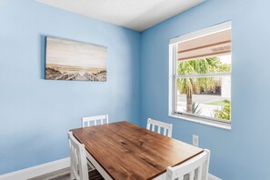A room with a wooden dining table, white chairs, a beach painting, and a window with a view of palm trees. - Spanish Villas #58 - Block From The Beach (Indian Rocks Beach)