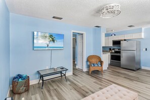 Una sala moderna con televisor de pantalla plana, una silla de mimbre y un comedor con cocina al fondo.