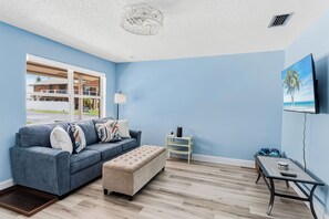 A living room with a blue wall, a grey sofa, a beige ottoman, a small white side table, and a wooden coffee table. - Spanish Villas #58 - Block From The Beach (Indian Rocks Beach)