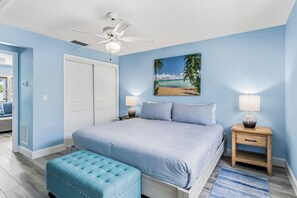 A bedroom with a bed, a ceiling fan, a painting of a beach, two bedside tables with lamps, and a blue upholstered bench. - Spanish Villas #58 - Block From The Beach (Indian Rocks Beach)