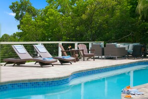 A poolside area with lounge chairs and a table, surrounded by trees. - The Beach Life - 4 BR 2.5 BA  premier home w/ pool & waterview (Indian Rocks Beach)