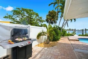 A barbecue grill with food, a pool, and palm trees. - The Beach Life - 4 BR 2.5 BA  premier home w/ pool & waterview (Indian Rocks Beach)