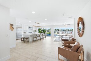A modern kitchen with white cabinets, a central island, and bar stools. There is a seating area with a brown sofa and chairs, and a view of a pool and buildings through large windows. - The Beach Life - 4 BR 2.5 BA  premier home w/ pool & waterview (Indian Rocks Beach)