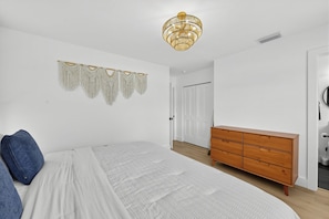 A bedroom with a bed, a wooden dresser, a macrame wall hanging, and a chandelier. - Central Tampa Oasis : Near Stadium, Downtown & TPA (Tampa)