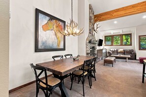 A dining area with a wooden table and chairs, a fireplace, and a large window with a view of trees. - Sage Haven | Walk to Ski + Hot Tub & Deck (Ketchum)