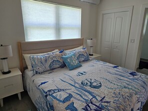 A bedroom with a bed, nightstands, a window with blinds, a door, and a wall-mounted air conditioner. - Emerald Escape - brand new 2-bedroom house on private beachfront property (Russell Island)