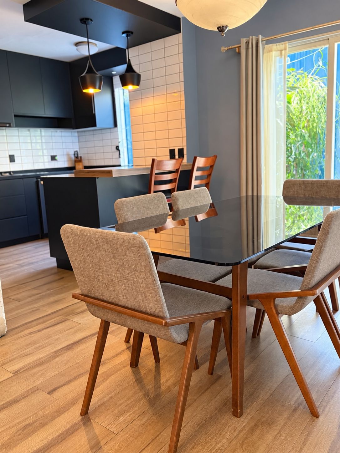 A modern kitchen with a dining area, featuring a glass table and wooden chairs, a black ceiling light, and a view of greenery outside.