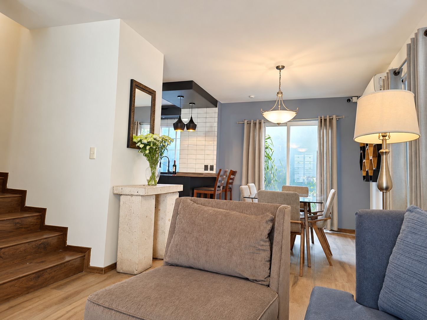A modern living room with a sofa, a dining area with chairs, a kitchen with a window, and a staircase.