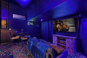 Ein Zimmer mit Spielbereich, inklusive einer Couch, Sesseln, einem Fernseher und einem leuchtenden Schild, das einen Spielraum anzeigt.