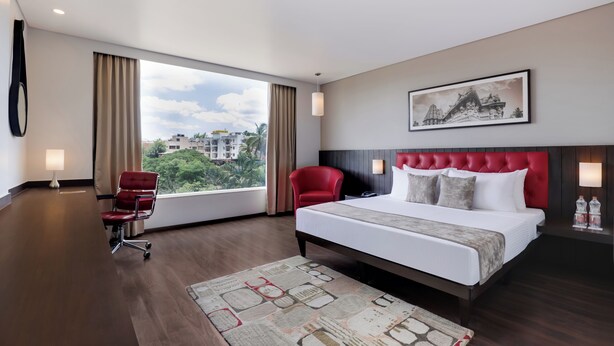 A hotel room with a large bed, a red chair, a desk, and a window with a city view. - The Phlox Hotel (Hubballi)