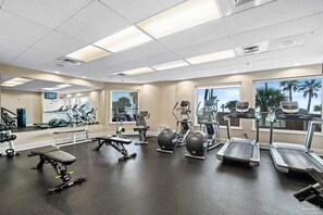 Ett gym med olika typer av träningsutrustning, stora fönster och utsikt över palmer.