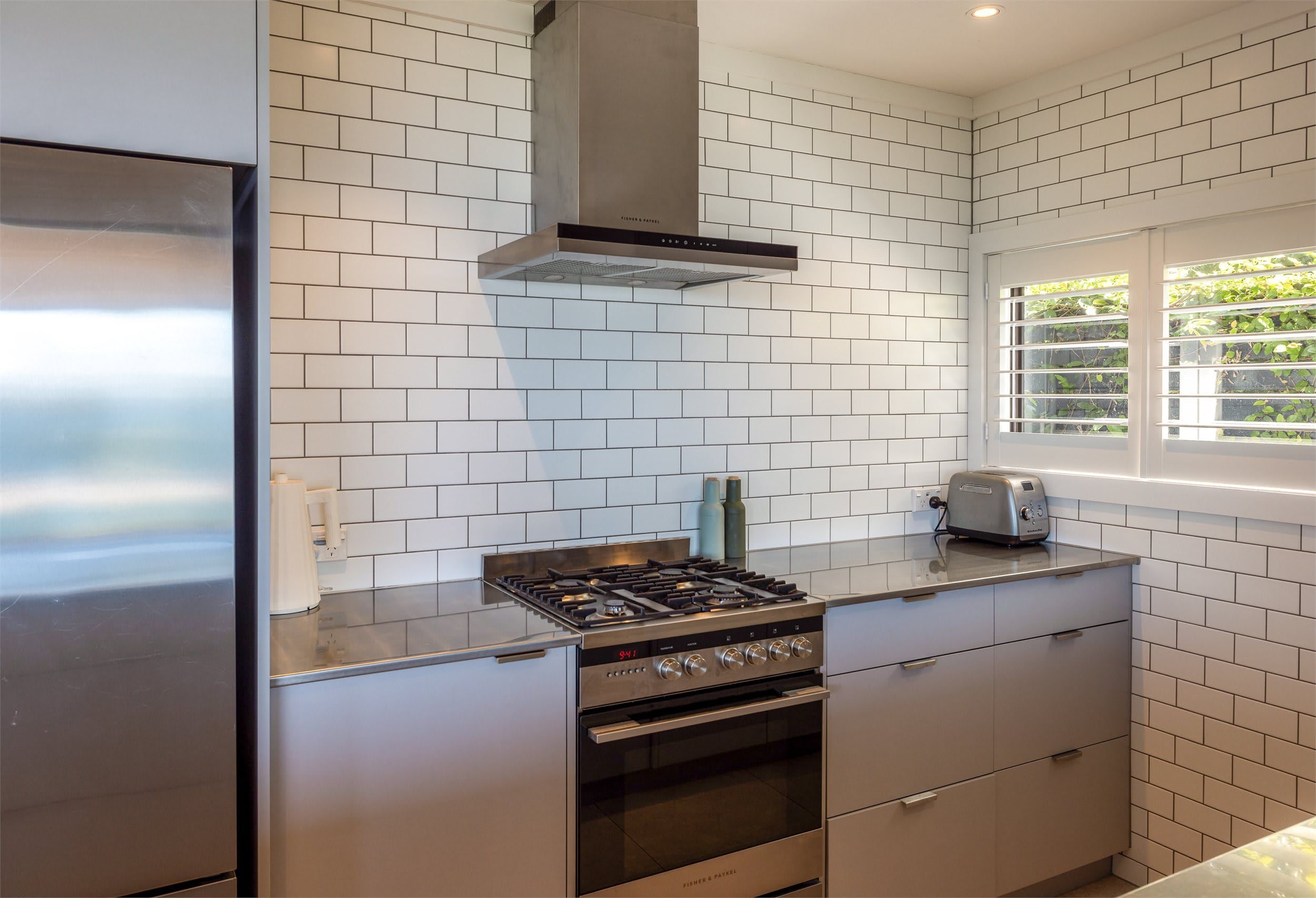 A kitchen with white tiled walls, a stainless steel range hood, a gas stove, an oven, and a toaster.
