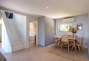 A modern kitchen with white cabinets, a dining area with wooden chairs, and a staircase leading to another level. - Sea Views & Space to Play (Waiheke Island)