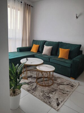 Ein Wohnzimmer mit einem grünen Sofa, zwei weißen runden Tischen, einem gemusterten Teppich und einem Blumentopf.