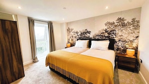A double bed with a yellow bedspread, two bedside lamps, a window with curtains, and a wall with a tree pattern. - 2BR Apartment with City Views and Parking (Leeds)