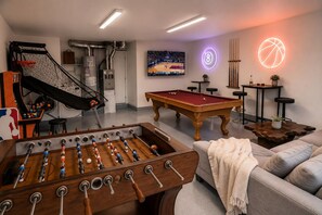 Una habitación con mesa de futbolín, mesa de billar, aro de baloncesto y un televisor mostrando un partido de baloncesto.