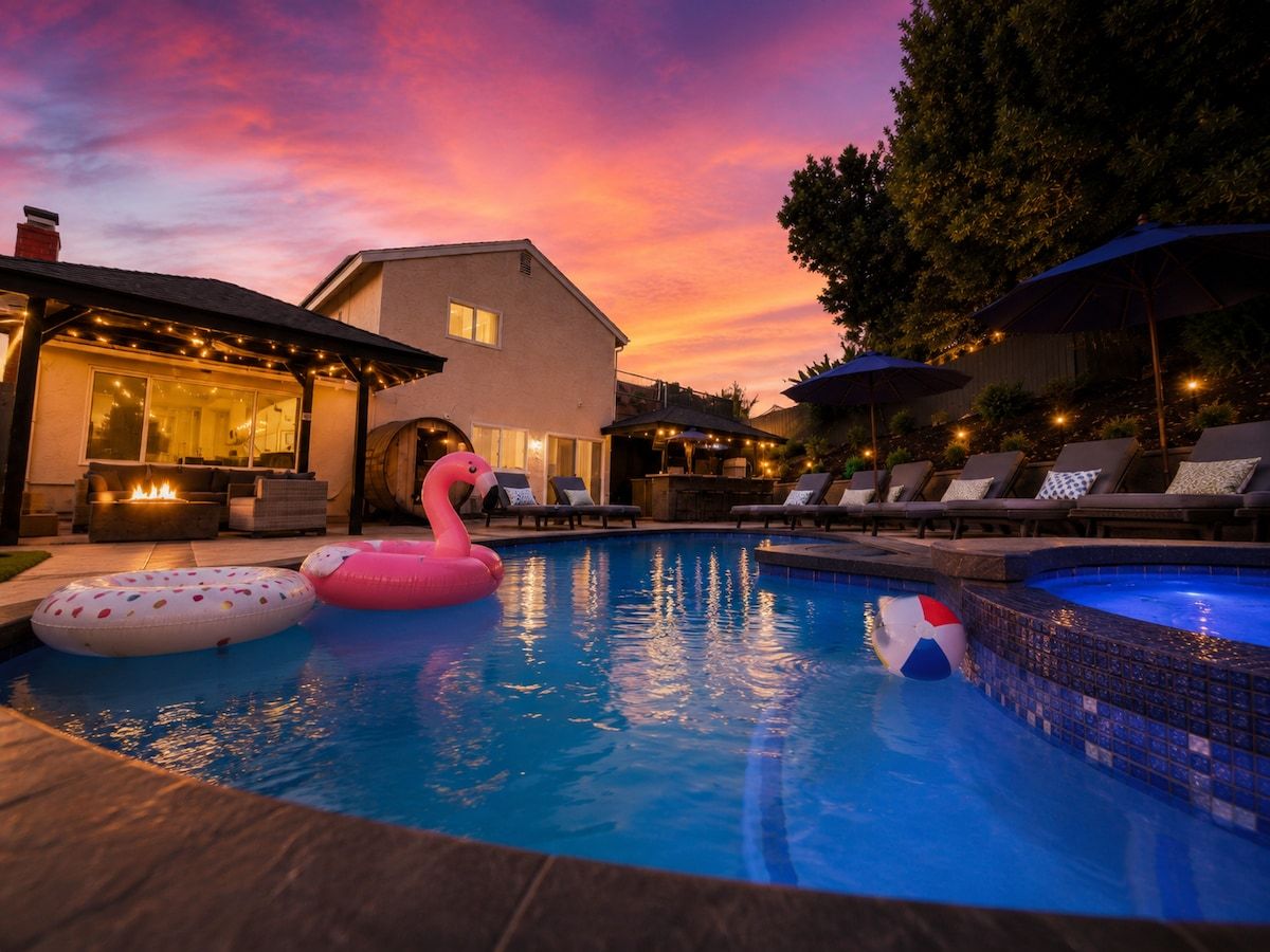 Una piscina con sillones de descanso y sombrillas, rodeada de una casa y árboles.