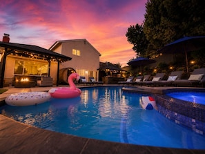 Una piscina con sillones de descanso y sombrillas, rodeada de una casa y árboles.