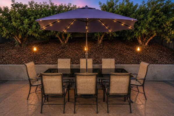 Un patio al aire libre con mesa y sillas, un paraguas grande y luces decorativas.