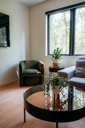 A modern living room with a leather armchair, a sofa, a round coffee table with a plant, and a large window with a view of trees. - NEW | Luxury Lakefront | Hot Tub, Beach & fishing (Backus)