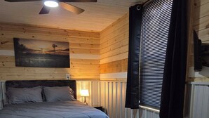 Quarto com cama, ventoinha no teto, mesa de cabeceira, abajur, janela com persianas e cômoda.