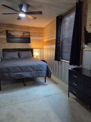 A bedroom with a bed, a ceiling fan, a nightstand, a lamp, a window with blinds, and a dresser. - The bunkhouse at Wilco Farms (Millen)