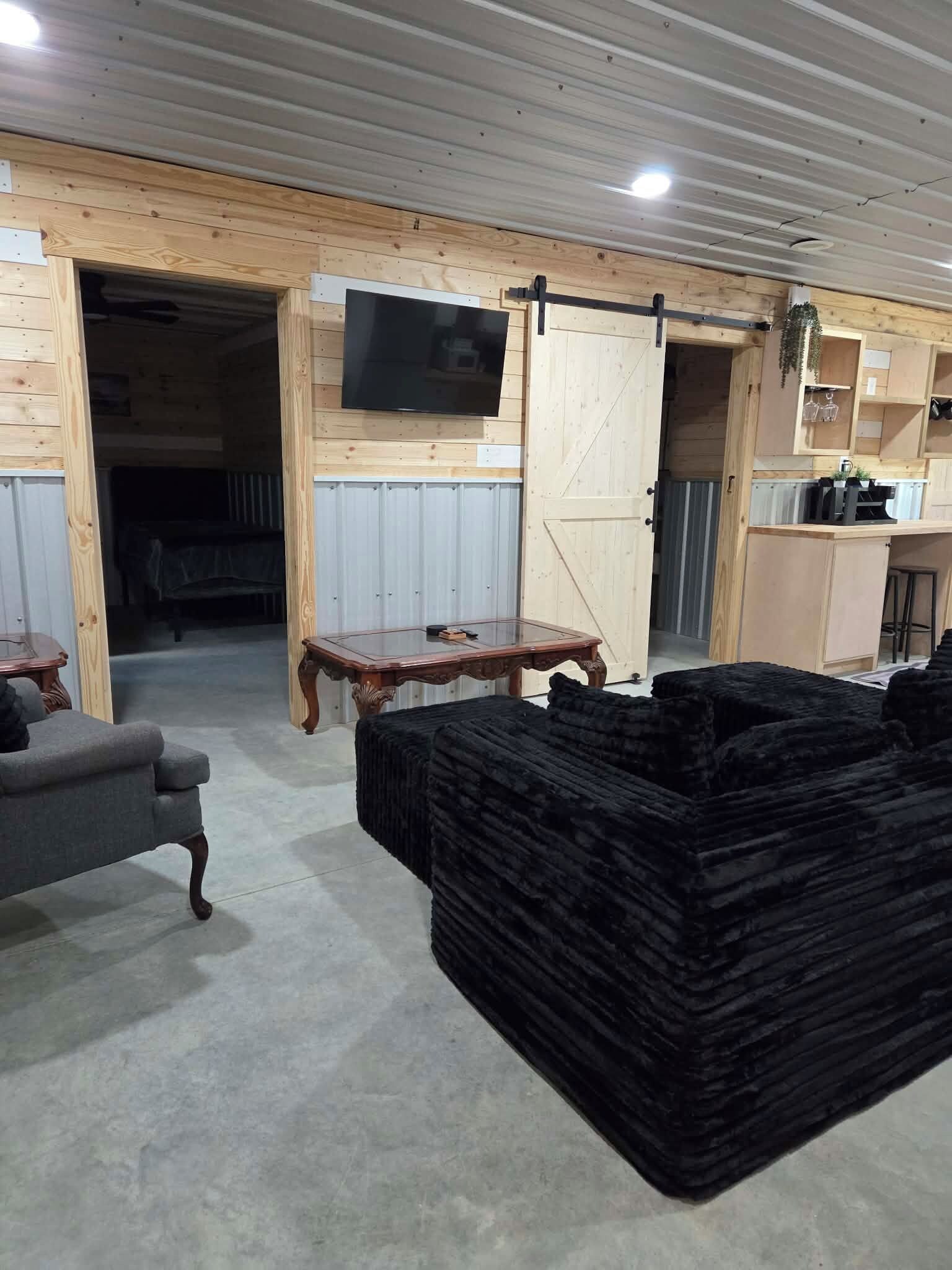 Quarto com decoração em madeira, televisão presa à parede, porta de celeiro deslizante, mesa de jantar e uma pilha de almofadas pretas.