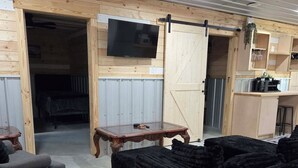 Quarto com decoração em madeira, televisão presa à parede, porta de celeiro deslizante, mesa de jantar e uma pilha de almofadas pretas.