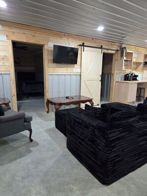 A room with a wooden interior, a television mounted on the wall, a sliding barn door, a dining table, and a stack of black cushions. - The bunkhouse at Wilco Farms (Millen)