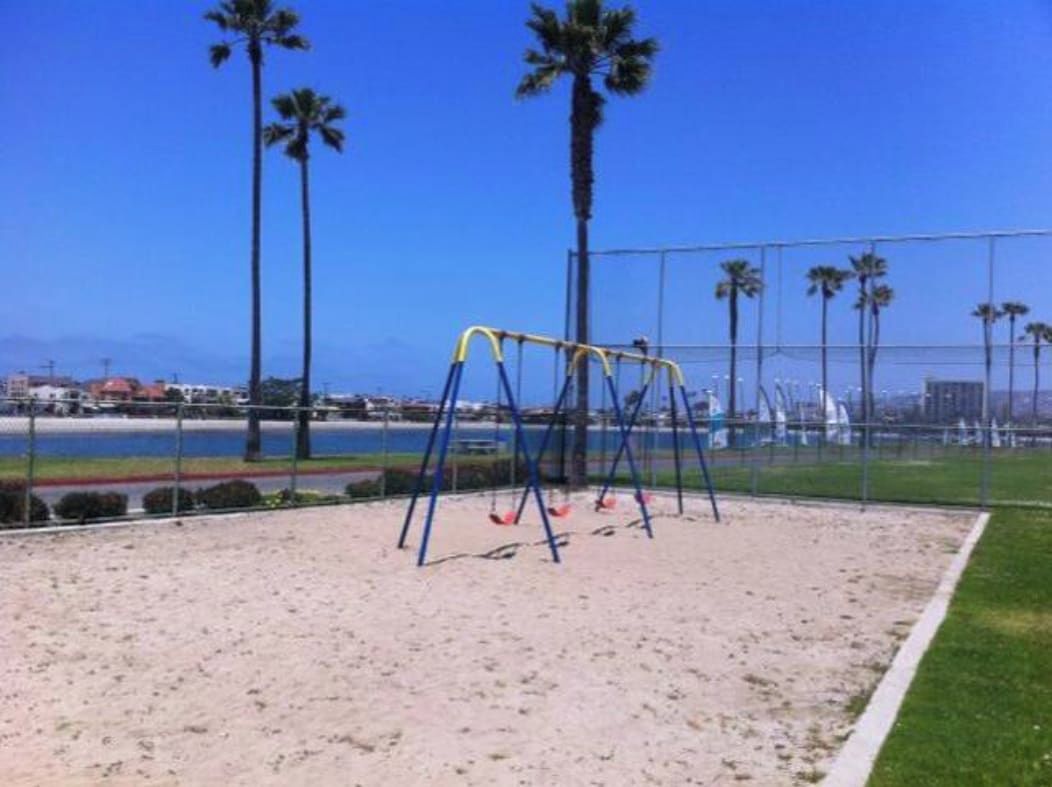 A sandy playground with swings and palm trees in the background.