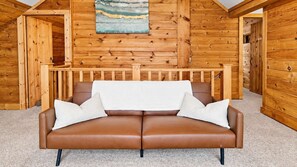 Een bruine leren bank met een witte plaid en kussens in een houten blokhutkamer met een schuine kap en een schilderij aan de muur.