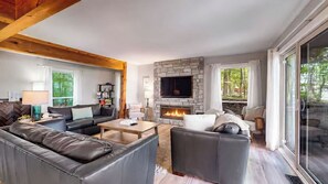 A modern living room with a stone fireplace, leather sofas, and a large window. - The Beach House | 6BR • Hot Tub • Steps to Lake! (South Haven)