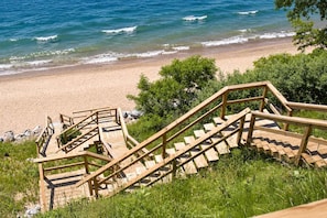 A wooden staircase leading to a beach with clear blue water. - The Beach House | 6BR • Hot Tub • Steps to Lake! (South Haven)