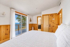 A bedroom with a large bed, wooden wardrobe, and a sliding glass door leading to a balcony with a view of the sea. - The Beach House | 6BR • Hot Tub • Steps to Lake! (South Haven)