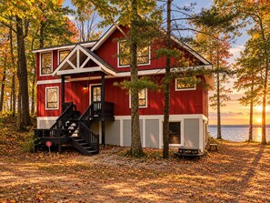 A red cabin with a staircase, surrounded by trees and a body of water. - The Beach House | 6BR • Hot Tub • Steps to Lake! (South Haven)