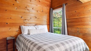 A wooden cabin room with a bed, a window, and a view of trees. - The Beach House | 6BR • Hot Tub • Steps to Lake! (South Haven)