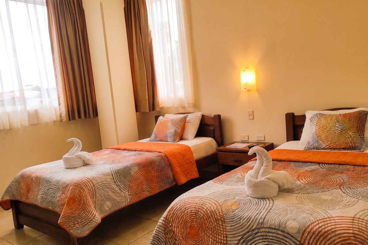 Habitación de hotel con dos camas, cada una con un cojín con forma de cisne, una mesita de noche y una lámpara de pared.
