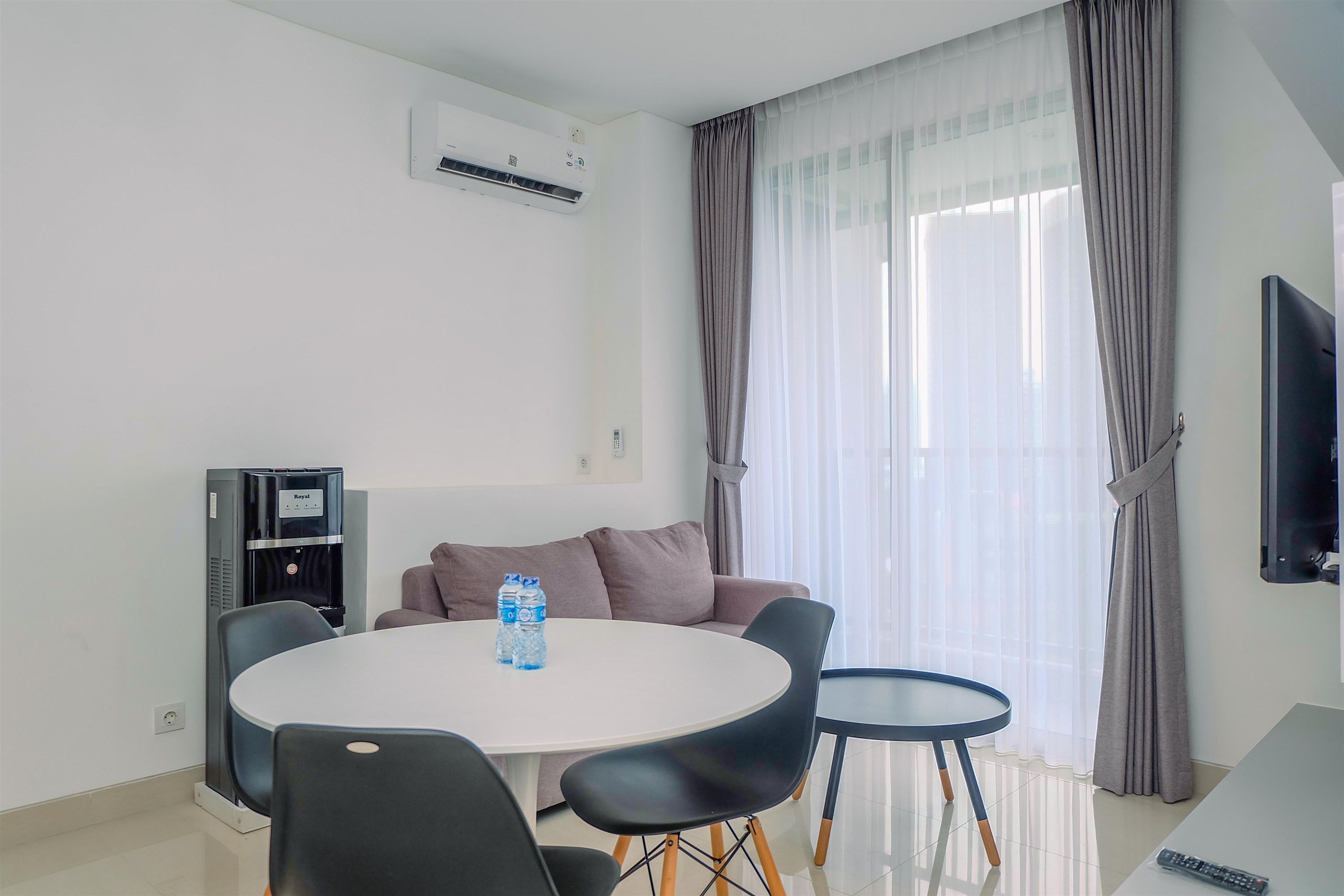 A modern living room with a round dining table, two chairs, a sofa, a water dispenser, and a flat-screen TV.