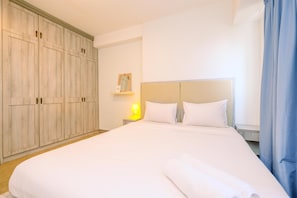 Quarto de hotel com um guarda-roupa grande, uma cama com lençóis brancos, uma mesa de cabeceira com abajur e uma janela com cortinas.