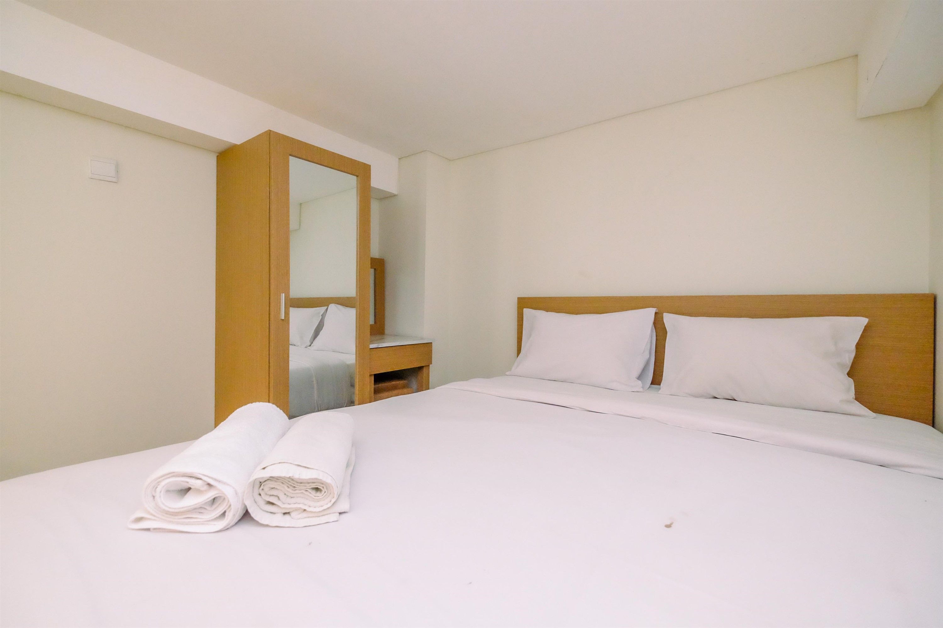 A hotel room with a bed, a wooden headboard, a nightstand, a mirror, and two rolled towels.